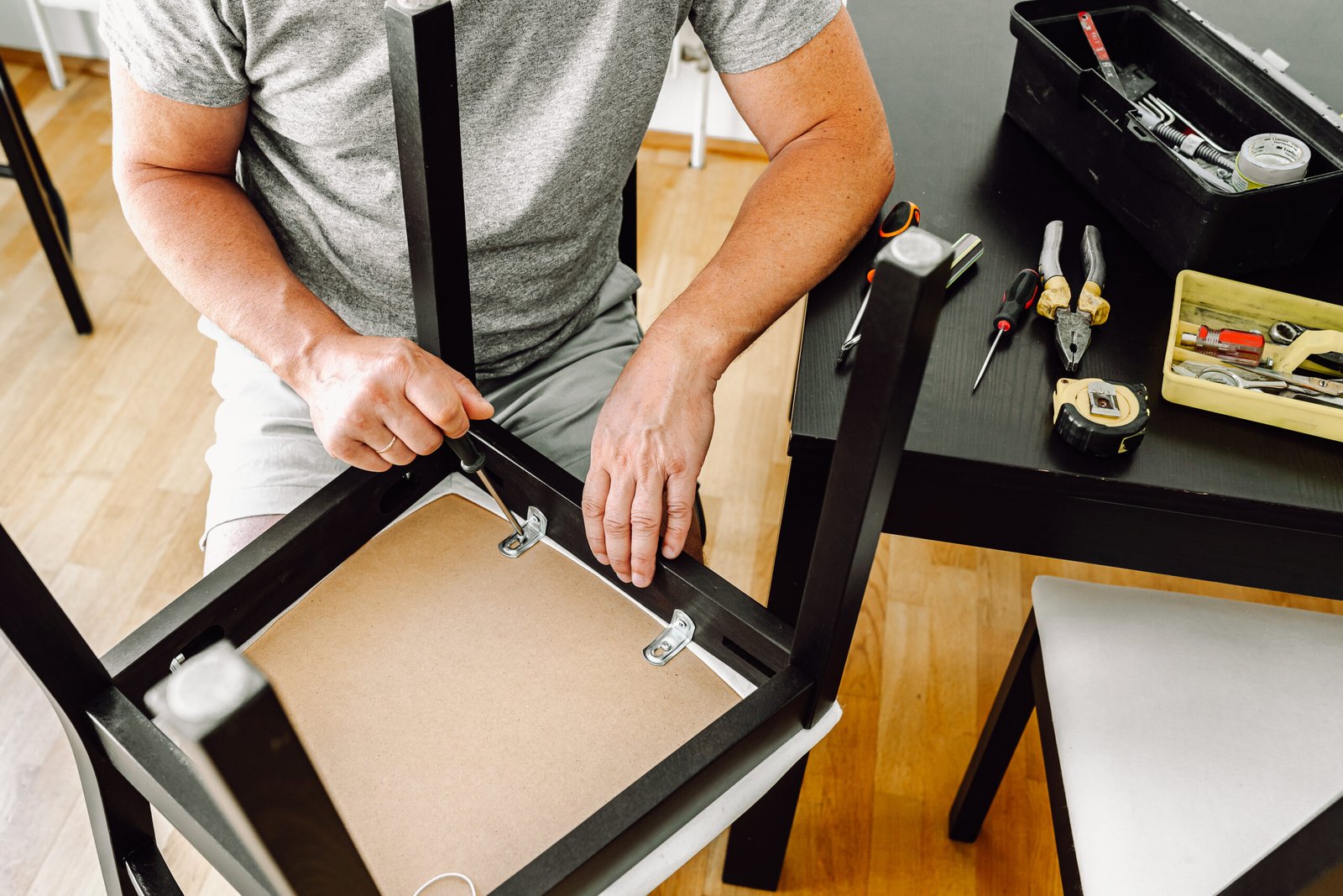 middle-aged man, gray-haired, muscular, attractive, repairs chair, tightens screw with screwdriver in wooden home chair. Repair home furniture, master carpenter, housework
