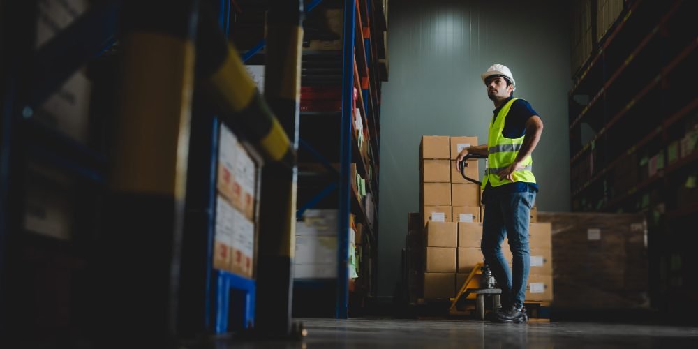 Handsome young male employee concentrating and focusing on work wearing safety helmet and uniform in warehouse while standing between racks in aisle