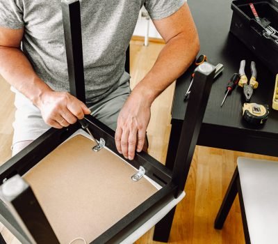 middle-aged man, gray-haired, muscular, attractive, repairs chair, tightens screw with screwdriver in wooden home chair. Repair home furniture, master carpenter, housework