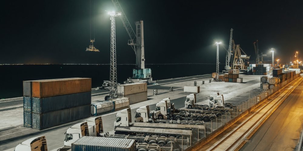 Industrial night scene at a seaport terminal with cargo trucks, shipping containers, and large cranes under bright artificial lighting, highlighting global logistics and freight transport operations